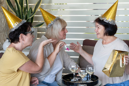 Happy senior women wearing party hats drinking wine, laughing, exchanging Christmas gifts, celebrating together at backyard patio. Middle aged woman with birthday hat sending blowing kiss.の写真素材