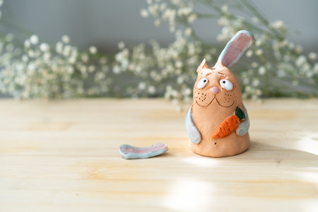 Clay Miniature Rabbit with Broken Ear Holds Carrot On Beautiful Flower Background. Handmade Sad Bunny with Broken Ear Looks at Camera.の写真素材