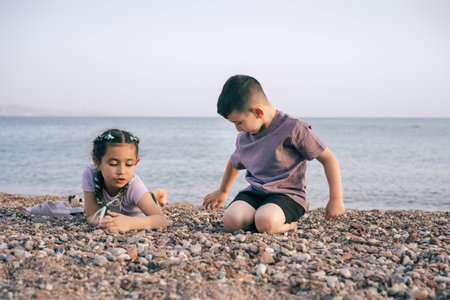 Children show each other sea pebbles. Two kids relaxing on sea beach in summer day. Siblings playing...