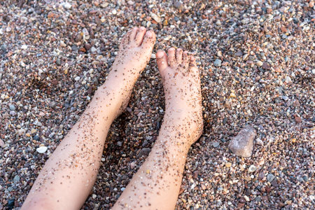 Two Feet In Sand On Beach Closeup. Child Legs On The Beach Full Of Small Pebbles On A Summer Day.