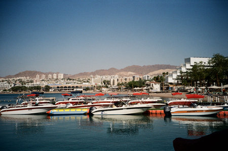 Eilat Israel, May 12, 2023. Waterfront And Mountain Backdrop. An analog photo capturing Eilats bustling waterfront with boats and distant hotels.のeditorial素材