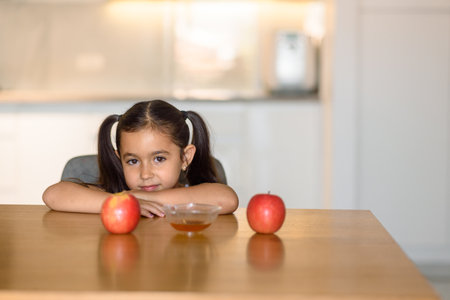 In a modern kitchen, a girl with pigtails is sitting at a table with apples and honey, smiling and appearing content.の写真素材