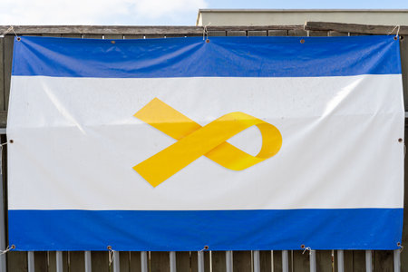 Close-up of a blue and white flag featuring a prominent yellow ribbon symbol, displayed on a fence. The flag represents support and awareness for the hostages in Israel.の写真素材