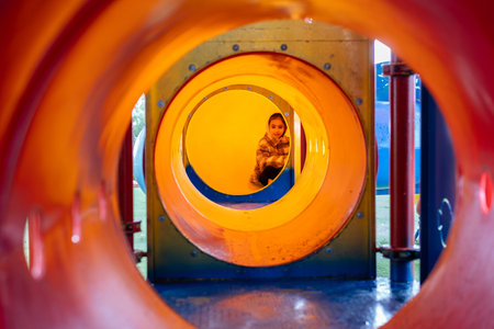 A child sits inside a vibrant orange playground tunnel, seen through circular openings, creating a unique layered perspective at the park on a rainy day.の写真素材