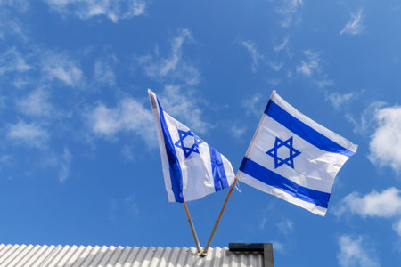 Two Israeli flags flying high above a rooftop, their movement reflecting strength and unity under a...