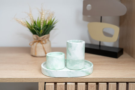 Two elegant concrete candle holders with a soft green hue are arranged on a round tray on a wooden table. A small plant in a tied fabric pot adds a touch of nature to the setting.の写真素材