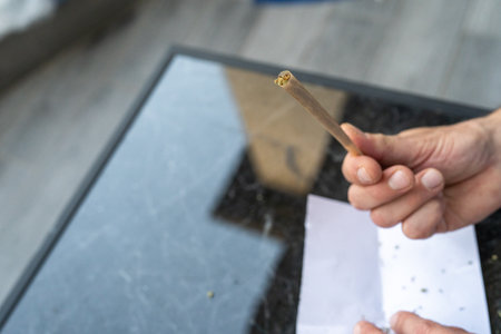 Man holds a cannabis joint above a dark marble table, with rolling papers and crushed herb nearby....