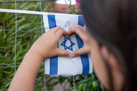 A child uses their hands to create a heart shape around an Israeli flag. The scene takes place...