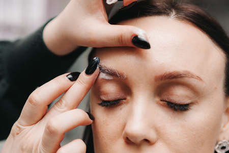 Close-up of a girl's face. The master holds a white pencil and draws the shape of the eyebrows for permanent makeupの写真素材