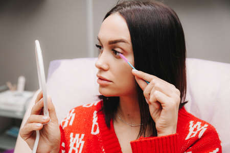 A beautiful girl looks in the mirror and applies makeup. the woman has increased her eyelashes and looks in the mirror.の写真素材