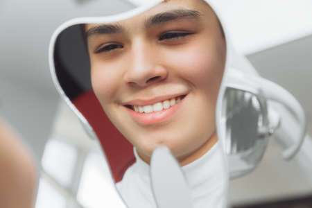 boy looks in the mirror with a toothy smile, sitting on a chair with a dentist in the dental office.の写真素材