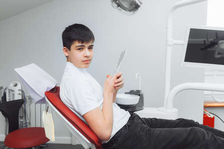boy looks in the mirror with a toothy smile, sitting on a chair with a dentist in the dental office.の写真素材