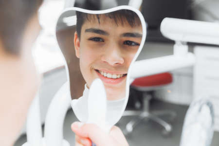 boy looks in the mirror with a toothy smile, sitting on a chair with a dentist in the dental office.の写真素材