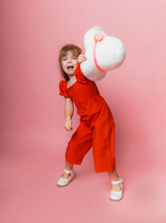 little cute girl in red overalls and white hat posing on a pink background.の写真素材