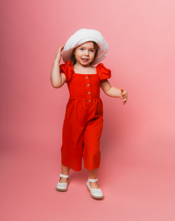little cute girl in red overalls and white hat posing on a pink background.の写真素材