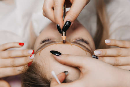 girl with permanent makeup on her eyebrows. The makeup artist makes markings with a brown pencil for eyebrow tattooing.の写真素材