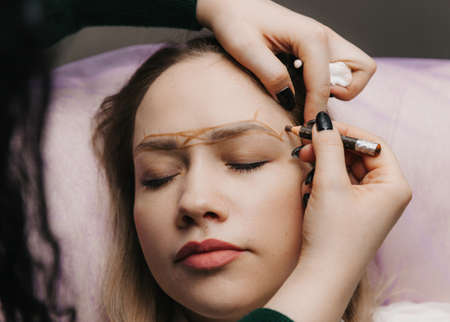 girl with permanent makeup on her eyebrows. The makeup artist makes markings with a brown pencil for eyebrow tattooing.の写真素材