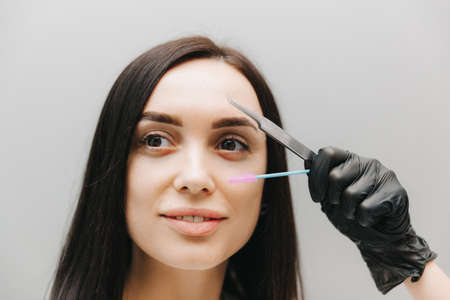 Close-up of a beautiful girl holding an eyebrow tweezer in her hand near her face in the studio