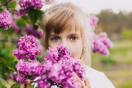 cute little girl in dress laughing in spring park near lilac. child smells lilac