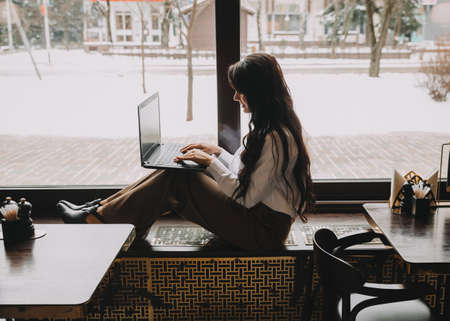 Confident young woman in a business suit works on a laptop while sitting by the window in a...