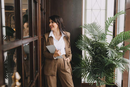 a woman in a business suit stands near the door in the office, smiling, holding a tablet