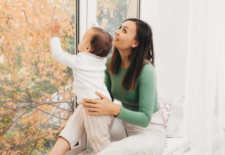 A young mother hugs her daughter, sitting in front of the window. Happy mom holding her baby while sitting on the windowsillの写真素材