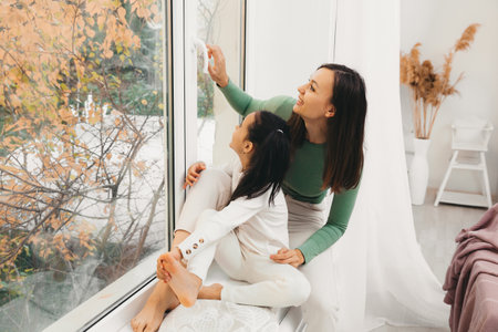 A young mother hugs her daughter, sitting in front of the window. Happy mom holding her baby while sitting on the windowsillの写真素材