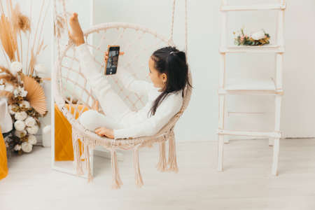 little girl is sitting and looking at her mobile phone, resting on a hanging chair.