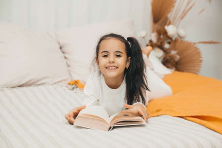 A little girl lies on the bed and reads a book. Bright bedroom interior.の写真素材