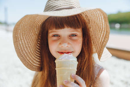 Charming little girl in a hat eats ice cream on the beach of the beach. Summer vacation concept.の写真素材