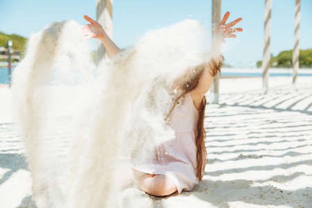 portrait of a little blonde girl in a dress sitting on the sand of the beach. Summer sunny day.