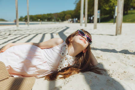 a little blonde girl in a straw hat lies on a sandy beach. girl sunbathing on the sandの写真素材