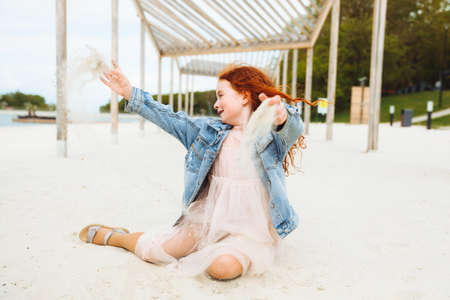 portrait of a little red-haired girl in a dress sitting on the sand of the beach. Summer sunny day.の写真素材