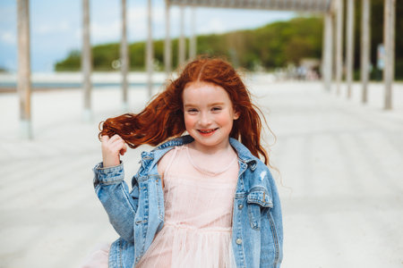 portrait of a little red-haired girl in a dress sitting on the sand of the beach. Summer sunny day.の写真素材