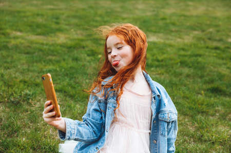 a little girl with red hair is holding a smartphone, taking a selfie. Video call concept.Modern...