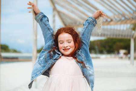portrait of a little red-haired girl in a dress sitting on the sand of the beach. Summer sunny day.の写真素材