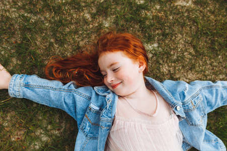 a little girl with red wavy hair lies on the grass in the park.