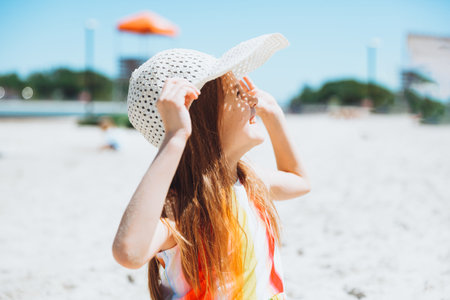 Portrait of a beautiful little girl in a striped dress and a straw hat sitting on the beachの写真素材