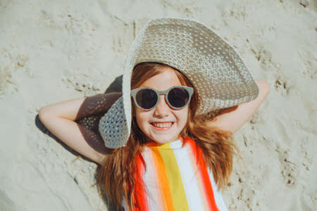 a little blonde girl in a straw hat lies on a sandy beach. girl sunbathing on the sandの写真素材