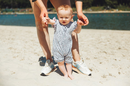 mother helps the girl to take the first steps on the beach. Lovely walking together on the sand on a summer day. Foreground.の写真素材
