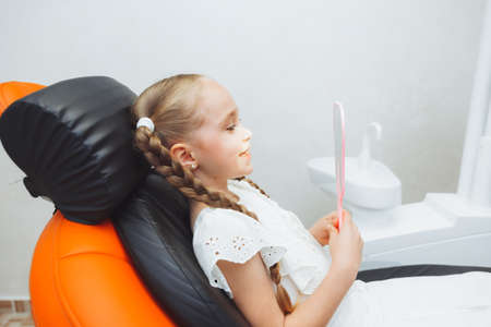 A little girl sits in a dental chair and examines her cured teeth while looking in the mirrorの写真素材