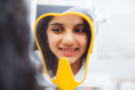A little girl sits in a dental chair and examines her cured teeth while looking in the mirrorの写真素材