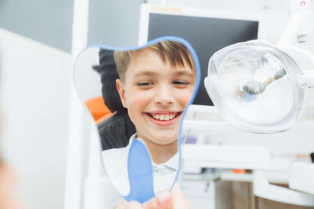 boy looks in the mirror with a toothy smile, sitting on a chair with a dentist in the dental office.