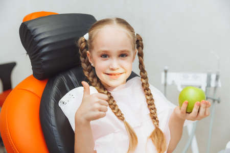 the girl at the reception at the pediatric dentist smiles and laughs. a little girl holds a big green apple in her hands. children's health concept.の写真素材
