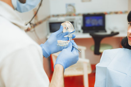 a male orthodontist dentist shows an artificial plastic jaw of a patient sitting in a dentist's...