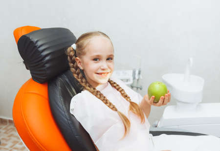 the girl at the reception at the pediatric dentist smiles and laughs. a little girl holds a big green apple in her hands. children's health concept.の写真素材