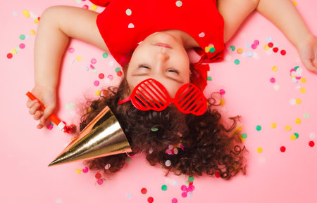 happy little girl in a birthday cap lies among the multicolored confetti on a pink background, a view from above.の写真素材