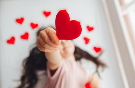 Valentine's Day, a little girl lying at home on a white bed among hearts, smiling and laughing with happiness, congratulating on the holidayの写真素材
