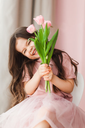 Portrait of a little girl with long dark hair close-up. The baby hugs a bouquet of fresh, delicate pink tulips. A gift for the holiday, spring time.の写真素材