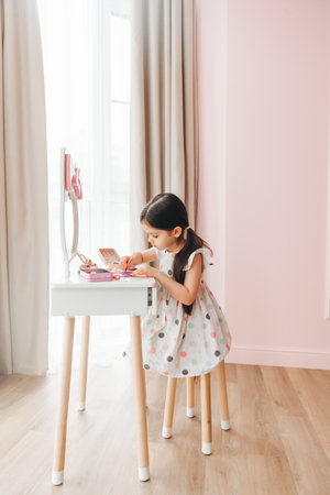 a beautiful little girl in the mirror preening. a little girl is sitting at a children's table and is wearing children's cosmetics.の写真素材